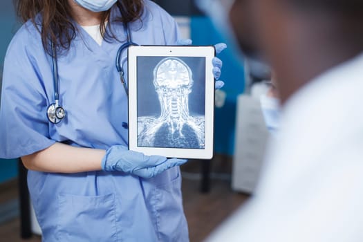 In medical office, doctor uses tablet to analyze a scan of a patient's skull. Selective focus of nurse in blue scrubs holding a device with an x-ray image. Advanced technology and expert care.