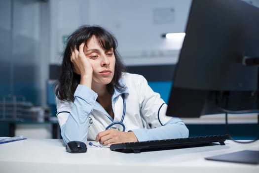 Worn out female doctor sitting at a desk with a desktop pc after a long day in hospital. Caucasian woman with her hand to her face, looking tired while doing healthcare research. Close-up shot.