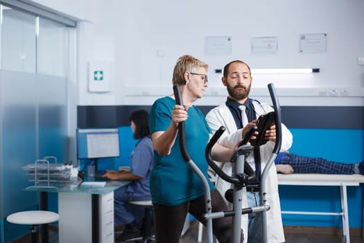 Doctor consults with old patient doing gymnastics for recovery at physiotherapy clinic. Aged woman uses stationary bicycle for physical exercise while talking to a healthcare specialist.