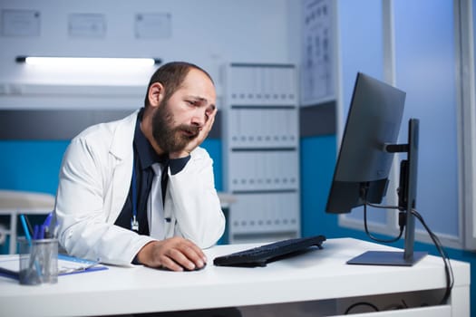 Bearded caucasian man wearing a lab coat, tiredly reviewing medical information on his desktop pc. Male doctor looking worn out is using computer in a hospital office room.