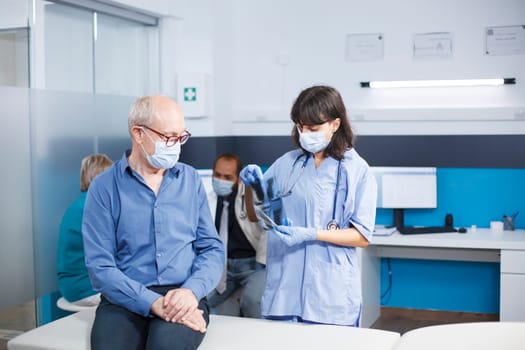 During coronavirus epidemic, nurse practitioner grasps CT scan data during medical consultation with elderly man. Healthcare professional describes radiography diagnostic and lung therapy.