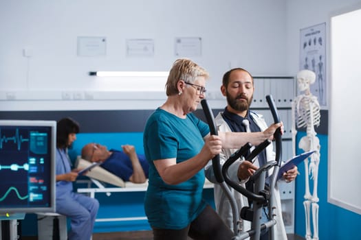 Medical specialist discussing physiotherapy with pensioner who is riding bike to heal from muscular injury. Elderly lady in physical treatment center using stationary bicycle for recovery.