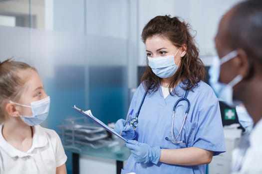 In medical office, female doctor wearing blue scrubs converses with young patient. Both individuals wear face masks. Medical equipment, including a stethoscope, is visible.