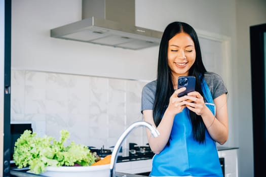 Cheerful woman leans on the kitchen table smiling while using her cellphone. Illustrating multitasking modern technology and a happy housewife cooking dinner at home.