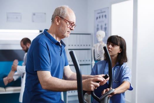 Female medical specialist assisting elderly outpatient on stationary bicycle for physical therapy. Senior man receiving physiotherapy to recover from injury with help from nurse at clinic.