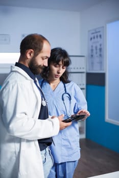 Male doctor and female nurse, together looking at patient files on digital tablet in the medical clinic office. Professional physicians using modern device that holds healthcare information.