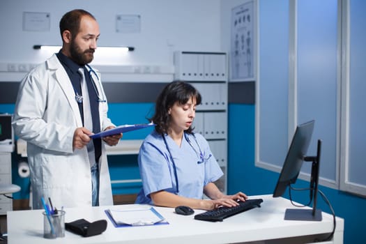 Caucasian doctor and healthcare worker collaborate in an office. They use technology, discuss medical charts, and wear scrubs and lab coats. Represents teamwork and communication in healthcare.