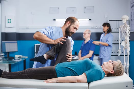 Chiropractor giving assistance to senior patient with knee injury for physical recovery. Woman doing leg exercise with help from medical assistant for osteopathic remedy and treatment.