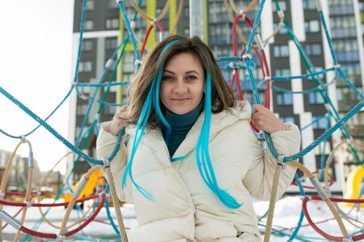 A woman with vibrant blue hair is sitting on a colorful playground structure. She looks relaxed as she enjoys the fresh air and the playful surroundings of the playground.