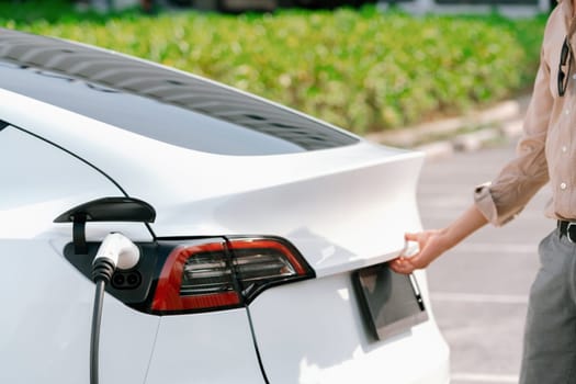 Young woman open trunk while EV car recharging battery from charging station in parking lot. Alternative energy and rechargeable electric vehicle for sustainable eco friendly travel. Expedient