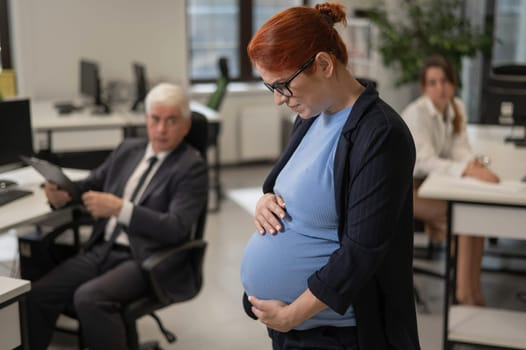 A pregnant woman suffers from pain and holds her stomach while standing in the middle of the office next to her colleagues