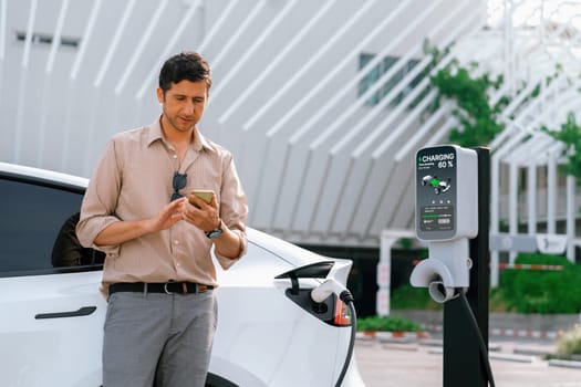 Young man use smartphone to pay for electricity at public EV car charging station at city commercial mall parking lot. Modern environmental and sustainable urban lifestyle with EV vehicle. Expedient