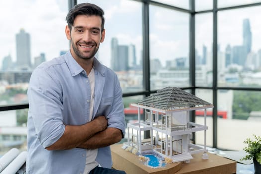 Portrait of architect engineer in casual outfit smile at camera while crossing arms. Businessman looking at camera and standing with arms folded near house model, architectural model. Tracery.