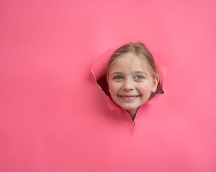 Cute Caucasian girl peeks out of a hole in a paper pink background