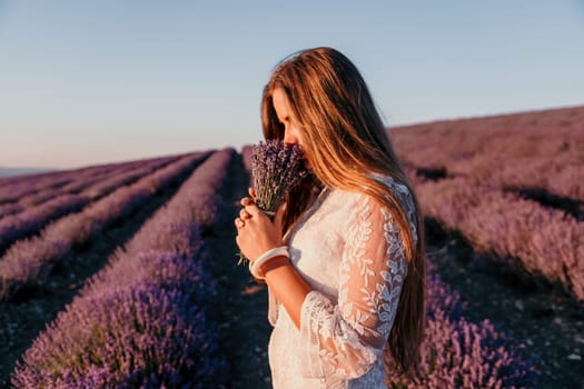 Close up portrait of young beautiful woman in a white dress and a hat is walking in the lavender field and smelling lavender bouquet.