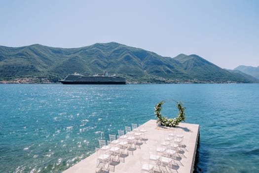 Rows of white chairs stand in front of a round wedding arch by the sea with a passing cruise ship in the background. High quality photo