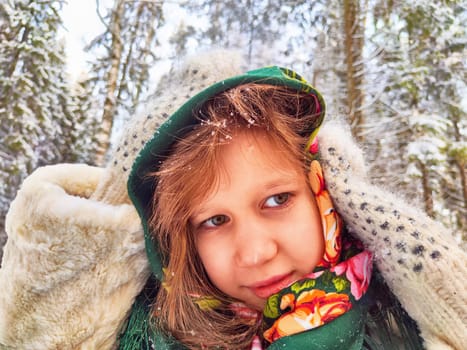 Cute girl in a traditional Russian headscarf and mittens on winter forest. Closeup portrait of child in folk clothes. Carnival Maslenitsa