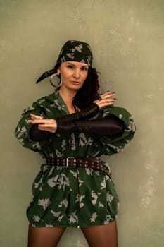 A woman in a green military uniform stands in front of a wall. She is wearing a bandana and has a confident posture