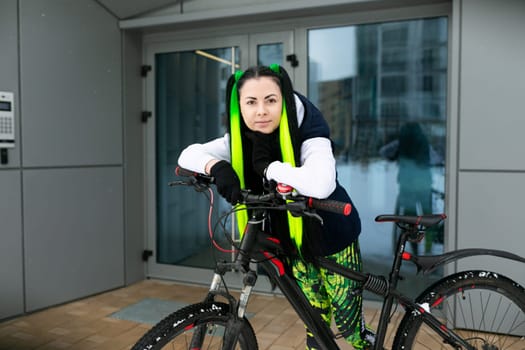 A woman wearing a black and green outfit is standing next to a bicycle. She is in a urban setting, looking confident and stylish.