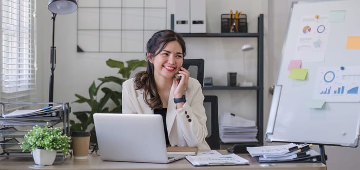 Young Asian business woman sits on the phone in an online business meeting using a laptop in a modern home office decorated with shady green plants..