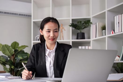 Young Asian business woman in a suit sits and reviews company work documents, checking information on a laptop on a work desk in a white office..