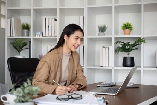 Young Asian business woman sits and works using a laptop in a modern office decorated with shady green plants..