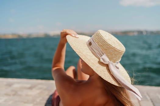 A woman in a swimsuit sits with her back holding a hat, looks at the ocean, sunny day, relaxes