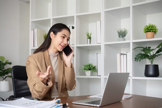 Young Asian business woman sits on the phone in an online business meeting using a laptop in a modern home office decorated with shady green plants..