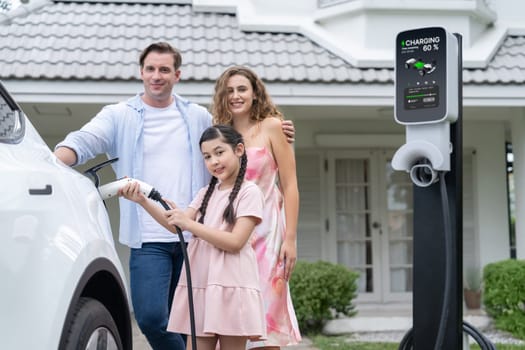 Happy little young girl learn about eco-friendly and energy sustainability as she help her family recharge electric vehicle from home EV charging station. EV car and modern family concept. Synchronos