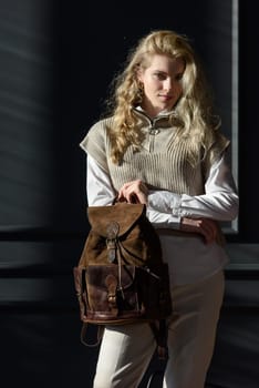 A woman holds brown leather backpack. Model wearing stylish knitted vest, white shirt and classic trousers.