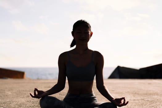 backlight of young woman doing meditation by the sea sitting with legs crossed, concept of mental relaxation and healthy lifestyle