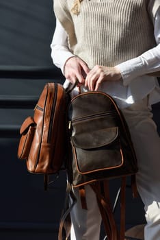 A woman holds two brown leather backpacks. Model wearing stylish knitted vest, white shirt and classic trousers.