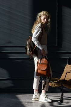 A woman holds two brown leather backpacks. Model wearing stylish knitted vest, white shirt and classic trousers.