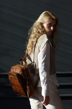 A woman holds brown leather backpack. Model wearing stylish knitted vest, white shirt and classic trousers.
