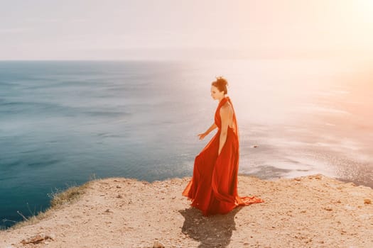 Side view a Young beautiful sensual woman in a red long dress posing on a rock high above the sea during sunrise. Girl on the nature on blue sky background. Fashion photo.