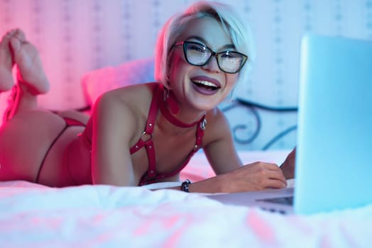 Sexy naked MILF girl in glasses and leather bdsm straps lies and communicates on the bed with laptop smiling emotionally erotically