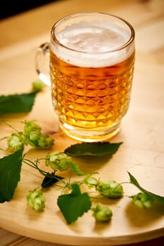 Large pint of beer with foam and sprigs of hops on bright wooden table