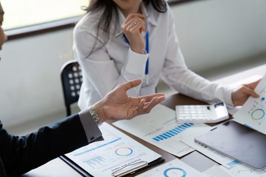 A man is talking to two women at a table with papers and a calculator. The man is wearing a watch and the women are wearing white shirts. The atmosphere seems to be professional