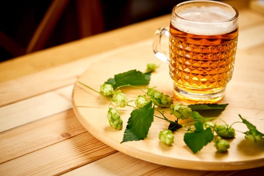 Large pint of beer with foam and sprigs of hops on bright wooden table