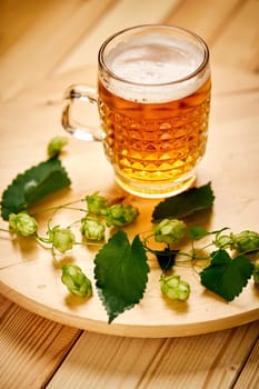 Large pint of beer with foam and sprigs of hops on bright wooden table