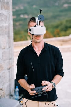 Young man in VR glasses with a joystick in his hands controls a drone while standing near a building. High quality photo