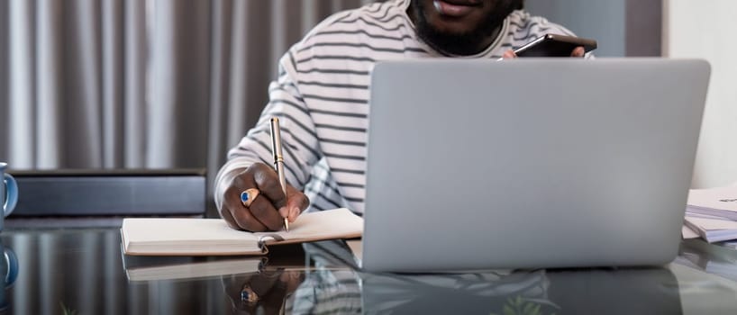 African American man working with laptop computer remote while sitting at glass table in living room. Black guy do freelance while taking notes work at home office.