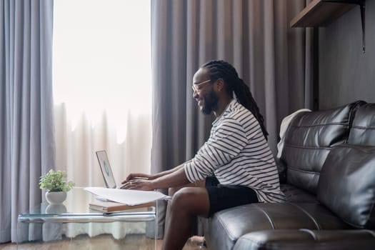 African American man working with laptop computer remote while sitting at sofa in living room. Black guy do freelance work at home office.