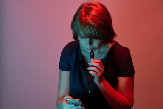 Beautiful attractive young woman with electronic cigarette staring at smartphone in neon light in studio