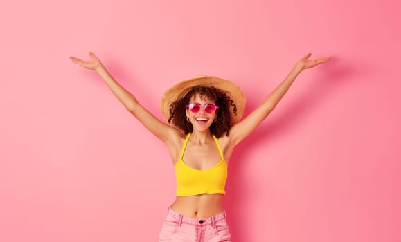 Happy cheerful African young woman in summer straw hat having fun raising her hands up on colorful pink studio background