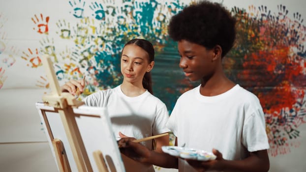 Diverse happy children paint canvas together with colorful stained wall. Smart multicultural student drawing together in art lesson while wearing white shirt. Creative activity concept. Edification.