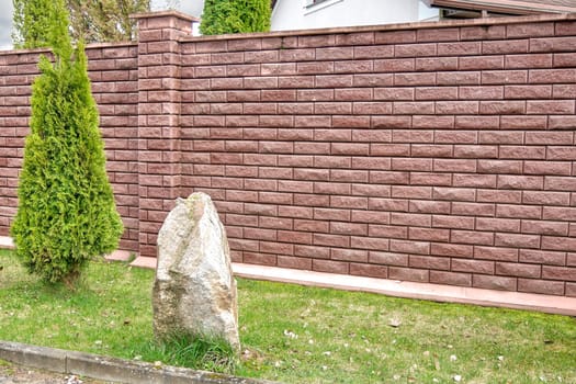 A red brick fence in front of the house next to a stone and a thuja tree
