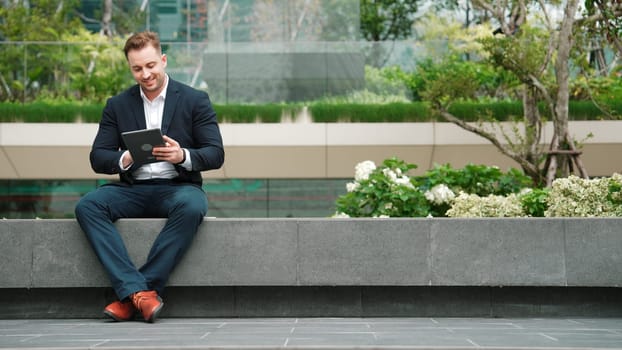 Caucasian smart businessman sitting at park while writing on tablet by using tablet pencil and planning marketing strategy. Manager looking at data analysis while thinking about financial plan. Urbane