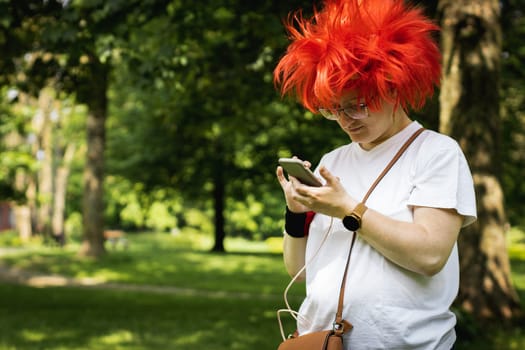 Portrait of a beautiful young caucasian woman in a red wig holding a smartphone in her hands stands in a city park on a sunny summer day, close-up side view.