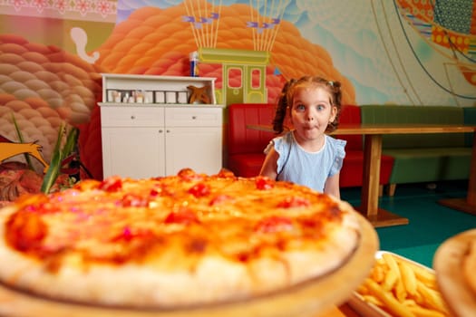 A young girl is seated in front of a large, delicious-looking pizza. She is gazing at the pizza, possibly deciding which slice to grab first. The table is set with plates and utensils, indicating that a meal is about to be enjoyed.
